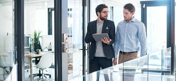 Two men in business attire, walking together. One man is holding a tablet device and pointing at it while the other man is looking at the tablet device.