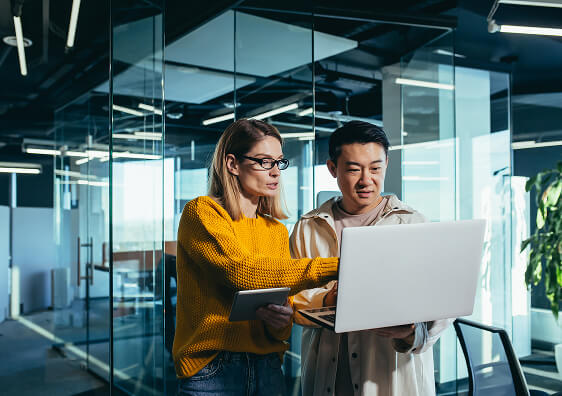 A man and woman in an office with glass walls. They are both looking at a laptop.
